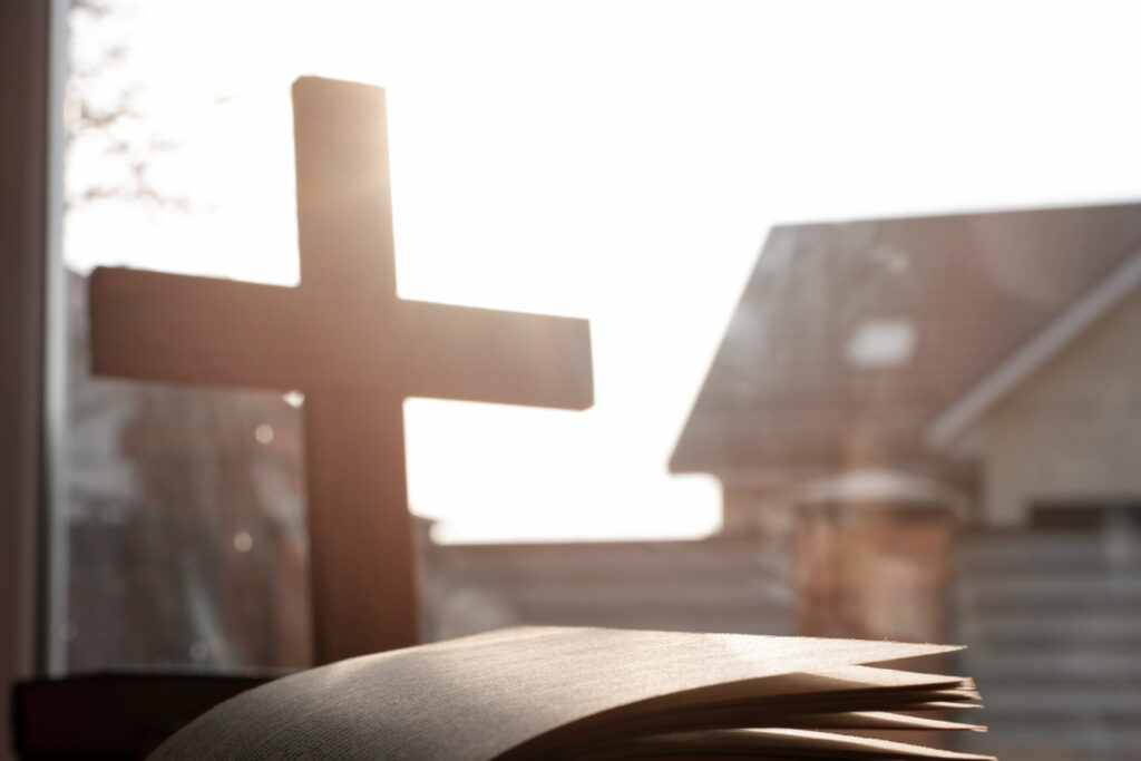 Bild von freepik Aufgeschlagene Bibel auf rotem Tuch, im Hintergrund ein Holzkreuz vor einem Fenster mit Blick auf Dächer. Licht fällt von außen ein.