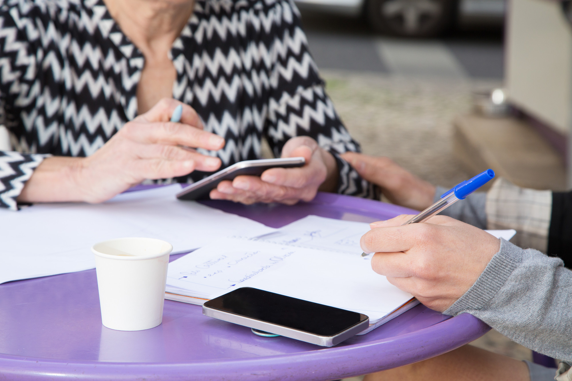 Zwei Personen sitzen an einem runden lila Tisch im Freien, schreiben in Notizblöcke und nutzen ein Smartphone. Auf dem Tisch liegen Papiere, Stifte, ein Becher und ein weiteres Smartphone.