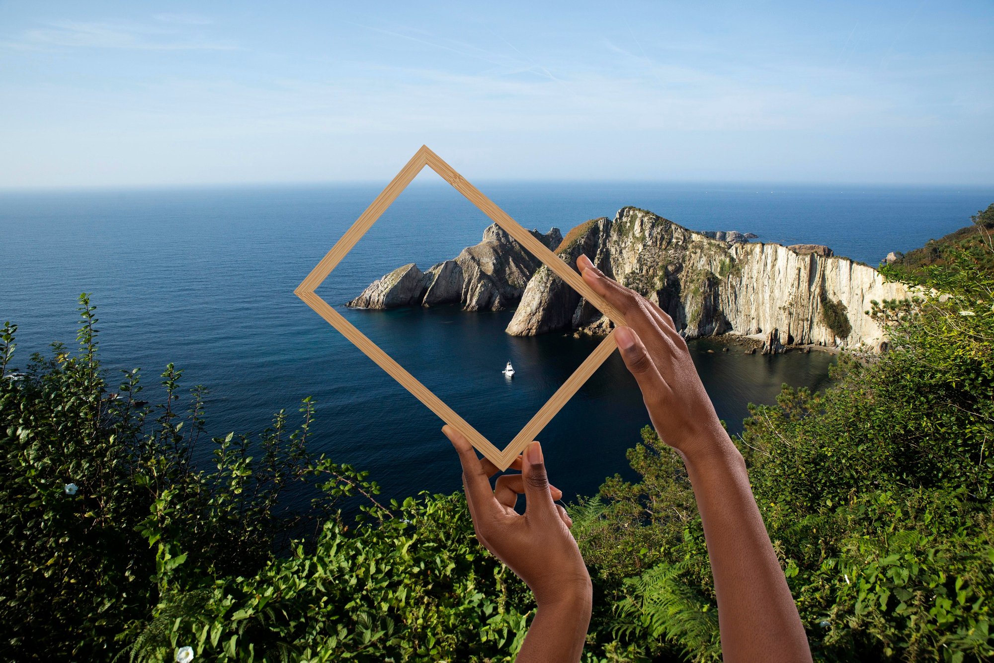Zwei Hände halten einen leeren Holzrahmen vor Felsküste am Meer. Ein Boot ist im Wasser sichtbar.