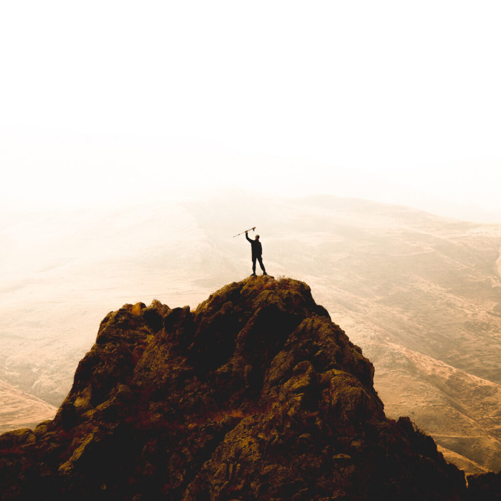 Einzelne Person steht auf einem felsigen Gipfel und hält einen Wanderstock in die Höhe. Weite Sicht in hellem Gegenlicht.