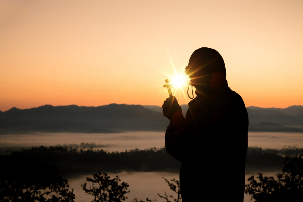 Silhouette einer Person mit einem Kreuz in der Hand. Sonnenaufgang hinter Berglandschaft im Hintergrund.