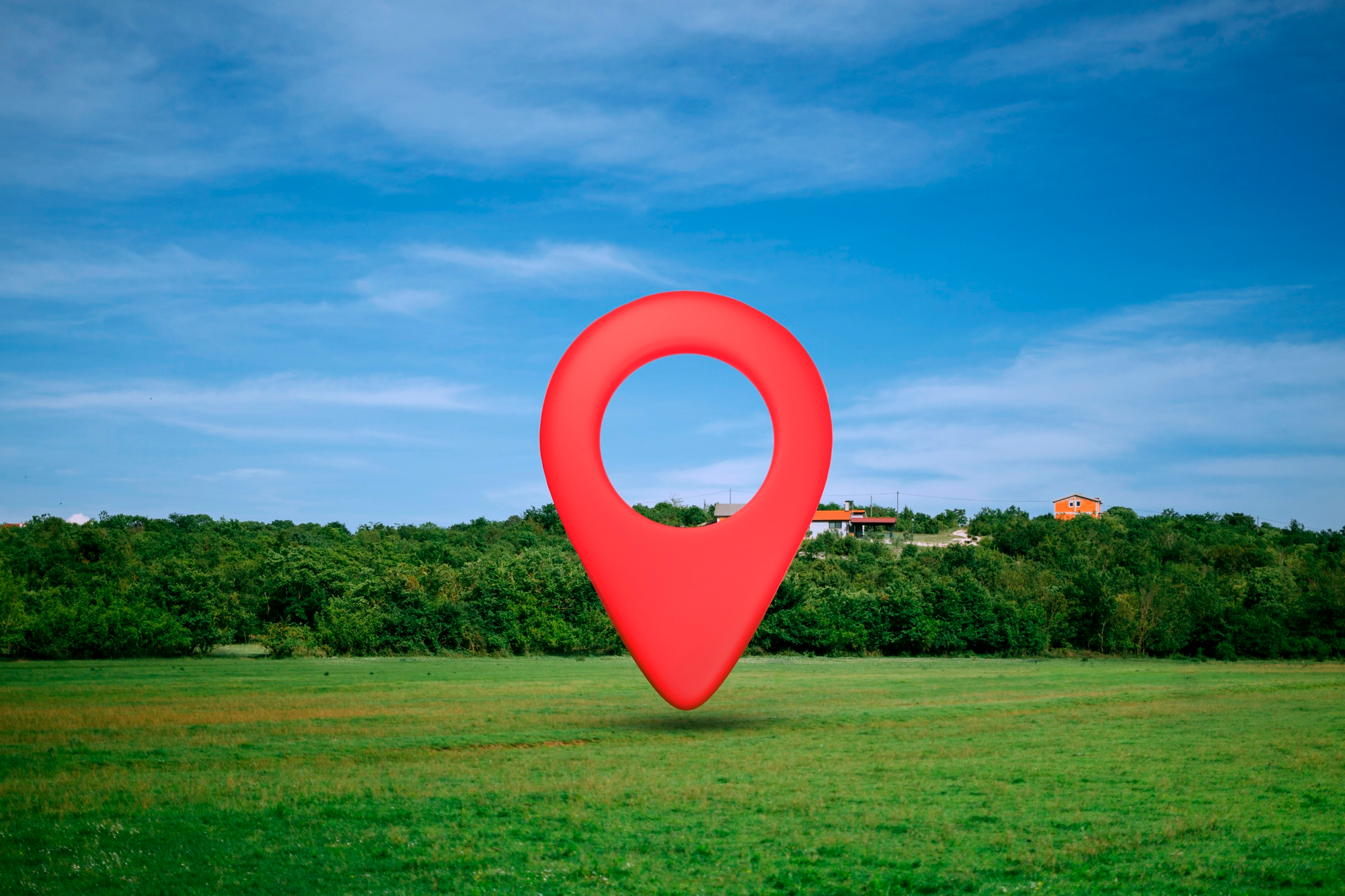 Ein großer roter Standortmarker steht auf einer grünen Wiese mit Bäumen im Hintergrund. Weiter hinten sind einzelne Häuser zu erkennen unter blauem Himmel.