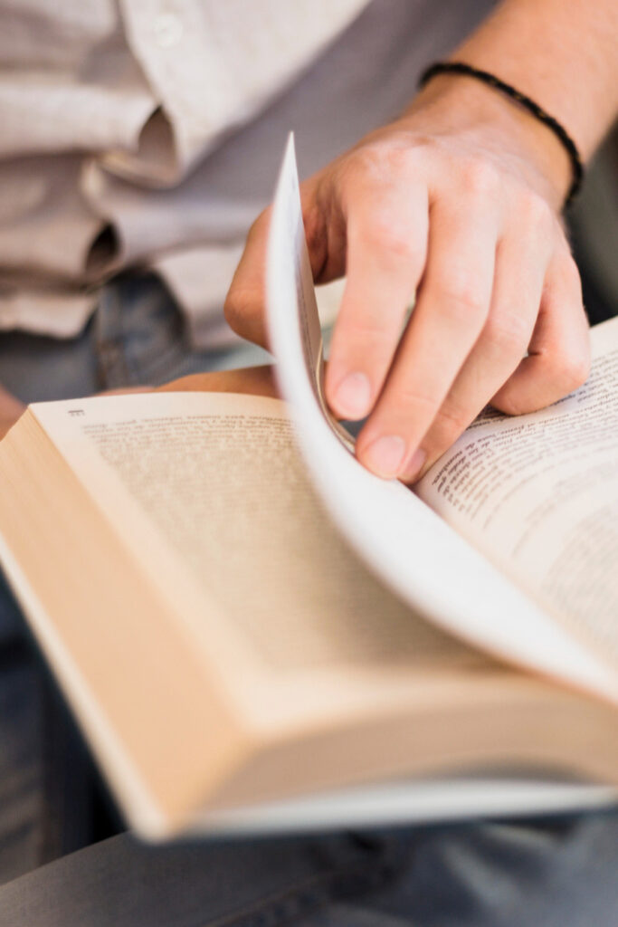Eine Person liest in einer Bibel und blättert um. Das Buch liegt auf dem Schoß, die Kleidung ist leger. Ein Armband ist sichtbar.