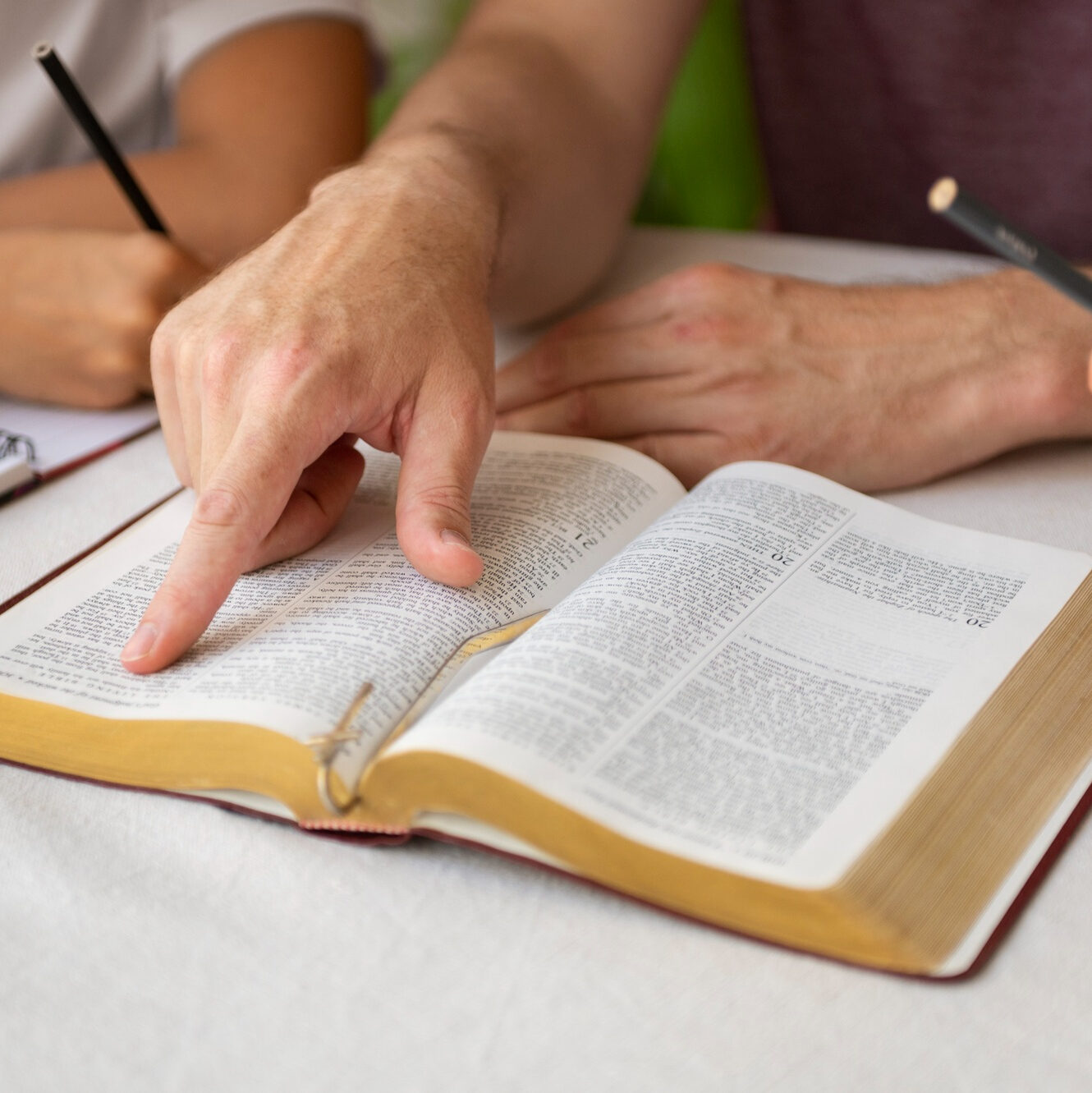 Zwei Personen an einem Tisch mit aufgeschlagener Bibel. Eine Hand zeigt auf eine bestimmte Bibelstelle, während andere Hände mit Bleistift erkennbar sind. Im Hintergrund liegt ein Notizbuch.