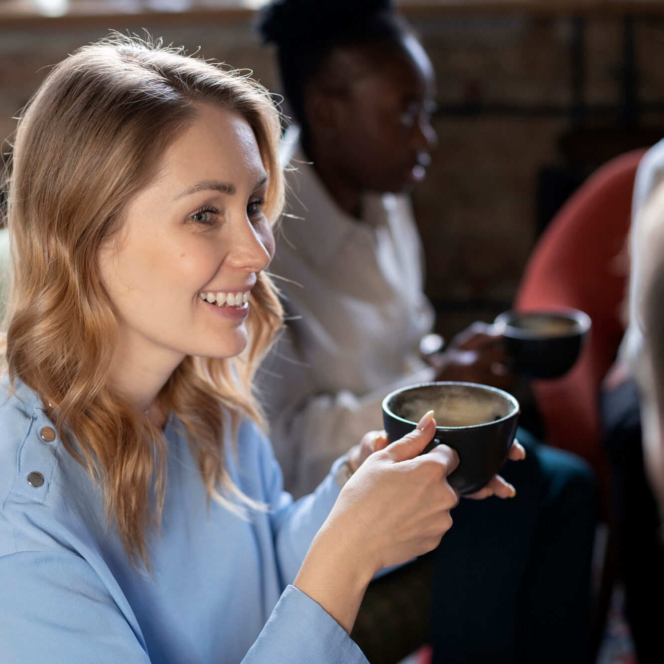 Blonde Frau in hellblauer Bluse hält eine schwarze Kaffeetasse und unterhält sich mit einer nicht vollständig sichtbaren Person. Im Hintergrund sitzt eine dunkelhäutige Frau ebenfalls mit Kaffeetasse in der Hand.