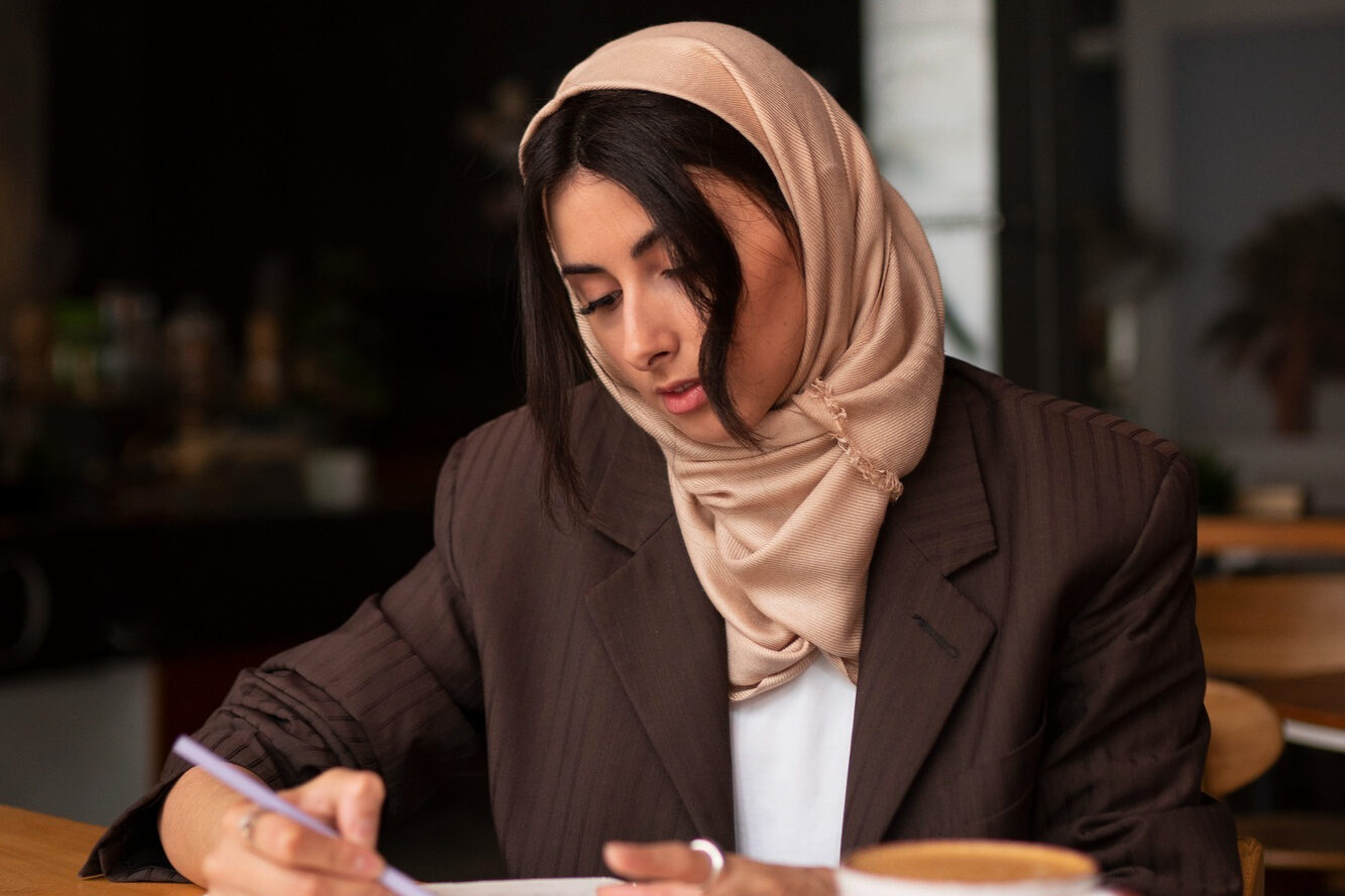 Frau mit beigefarbenem Kopftuch und dunklem Blazer sitzt an einem Tisch in einem Café und schreibt in ein Notizbuch, vor ihr steht eine Tasse.