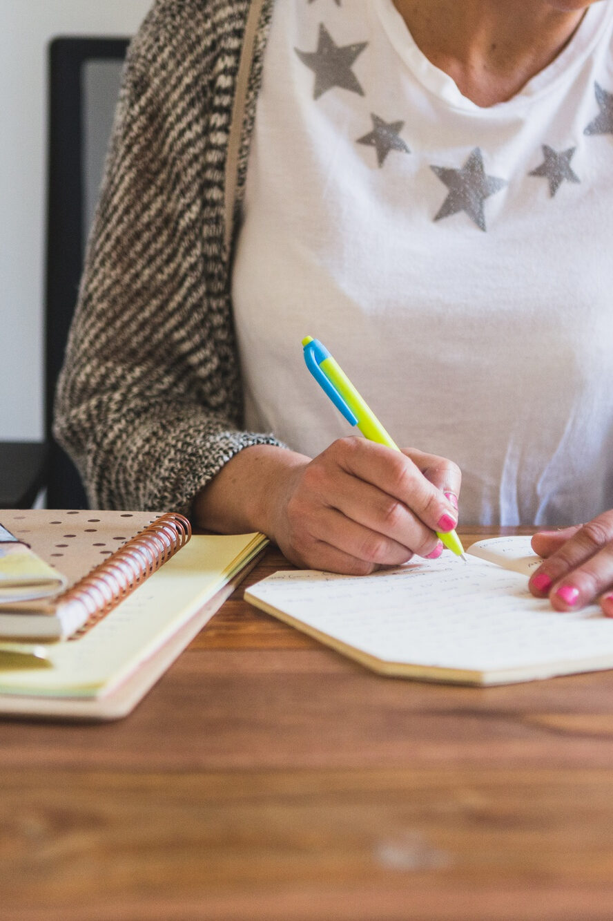 Frau mit Sternen-T-Shirt und Strickjacke sitzt am Tisch und schreibt mit einem gelb-blauen Stift in ein Notizbuch, daneben liegen weitere Hefte und Unterlagen.