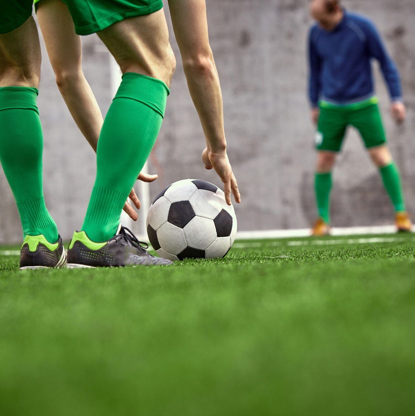 Ein Fußballspieler mit grünen Stutzen stellt den Ball auf dem Spielfeld ab. Im Hintergrund steht ein weiterer Spieler unscharf vor einer Betonwand.