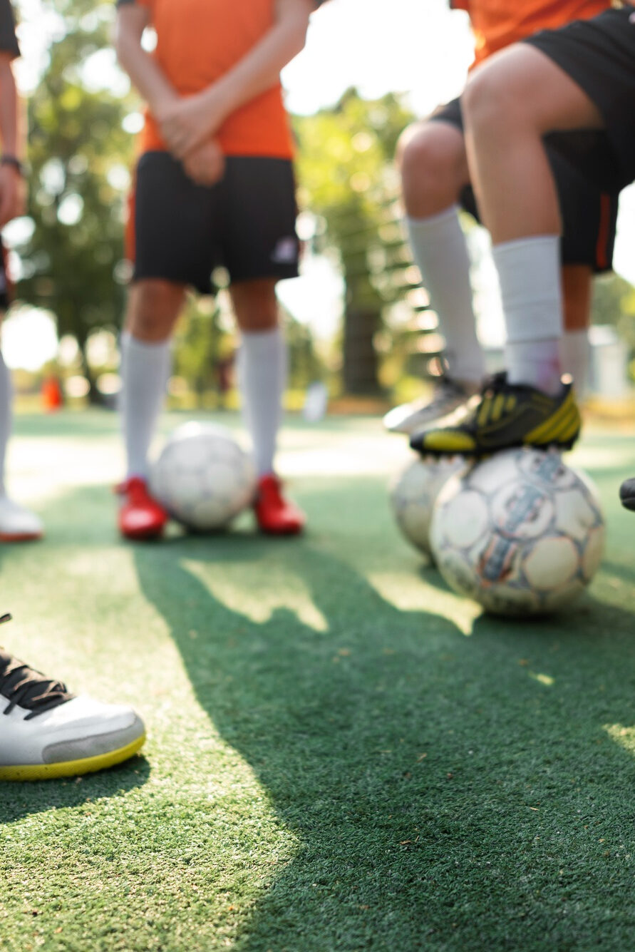 Gruppe von Fußballspielern in kurzer Trainingskleidung und weißen Stutzen steht im Kreis auf einem Kunstrasenplatz, mehrere Spieler haben einen Fuß auf einem Ball.