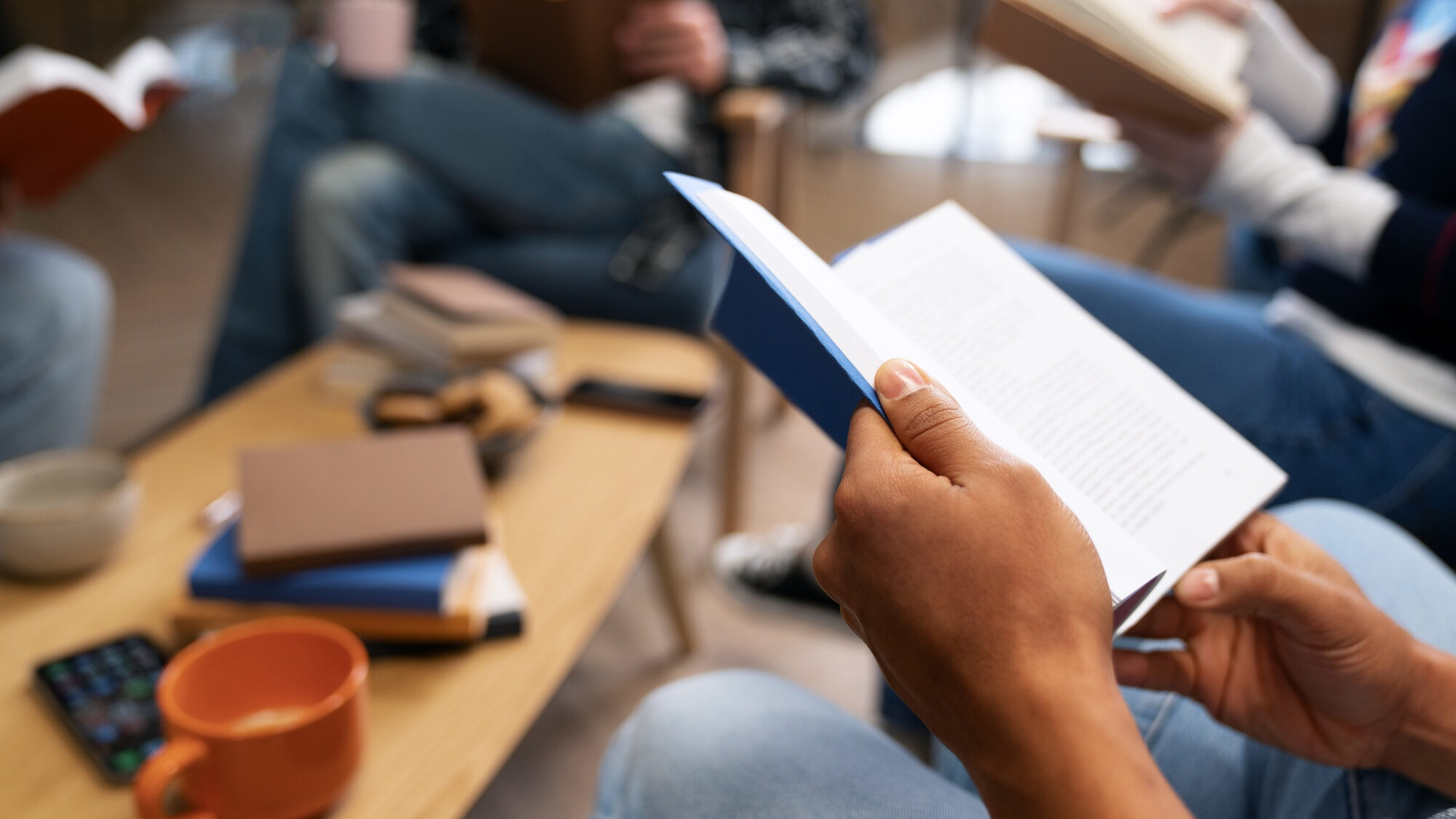 Mehrere Personen sitzen im Kreis und lesen gemeinsam in Bibeln. Im Vordergrund ist eine Hand mit aufgeschlagener Bibel sichtbar. Auf dem Tisch liegen weitere Bücher, Tassen und ein Smartphone.