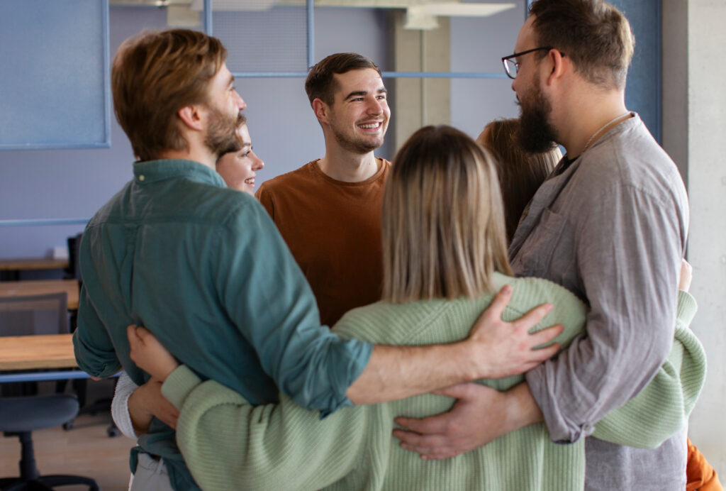 Sechs Personen stehen im Kreis und umarmen sich gegenseitig. Alle wirken fröhlich und blicken einander an.