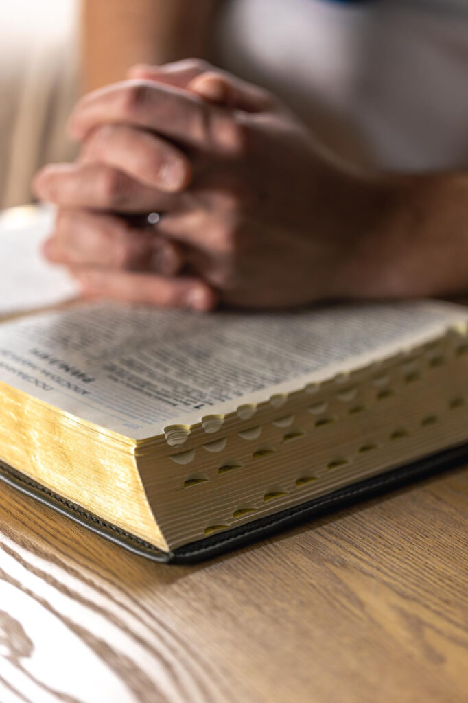 Bild von freepik Hände in betender Haltung liegen auf einer aufgeschlagenen Bibel mit Registerstanzung, die auf einem Holztisch liegt.
