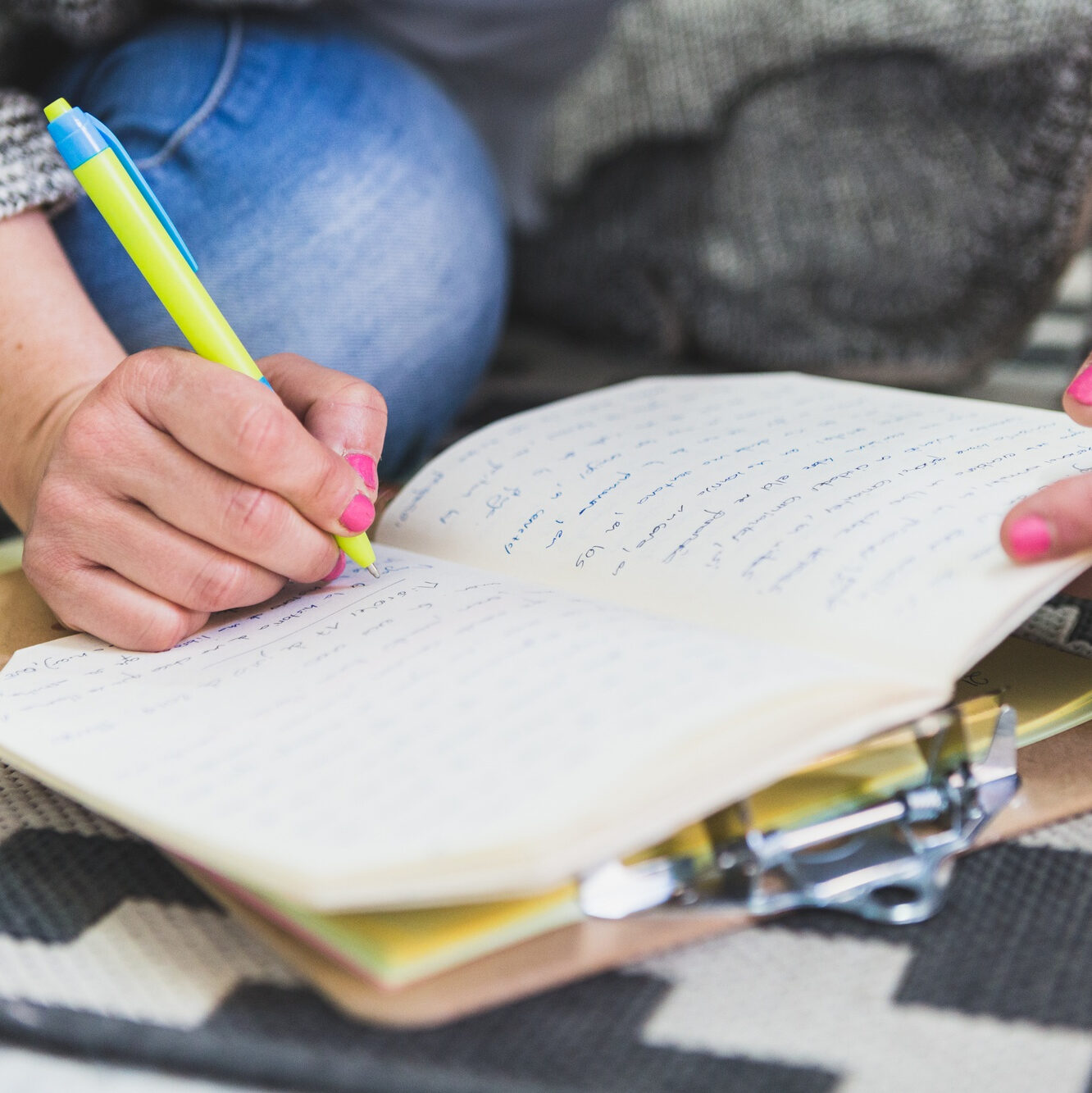 Hände mit pink lackierten Fingernägeln schreiben mit einem gelb-blauen Stift in ein Notizbuch auf einem Teppichboden, daneben eine Schreibmappe.