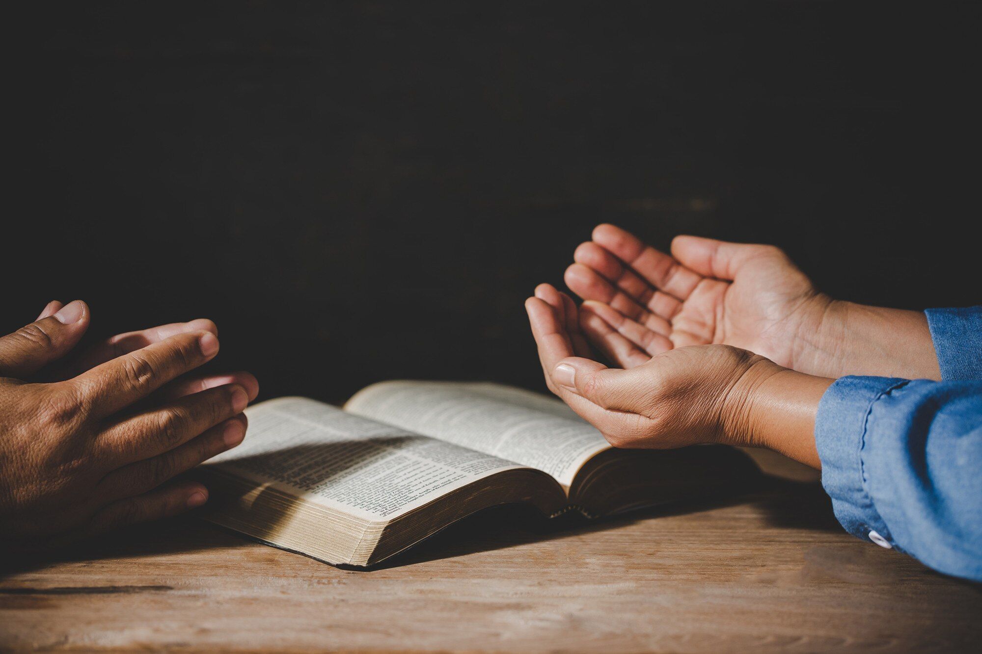 Zwei Paar Hände in Gebetshaltung vor einer aufgeschlagenen Bibel auf einem Holztisch, dunkler Hintergrund.