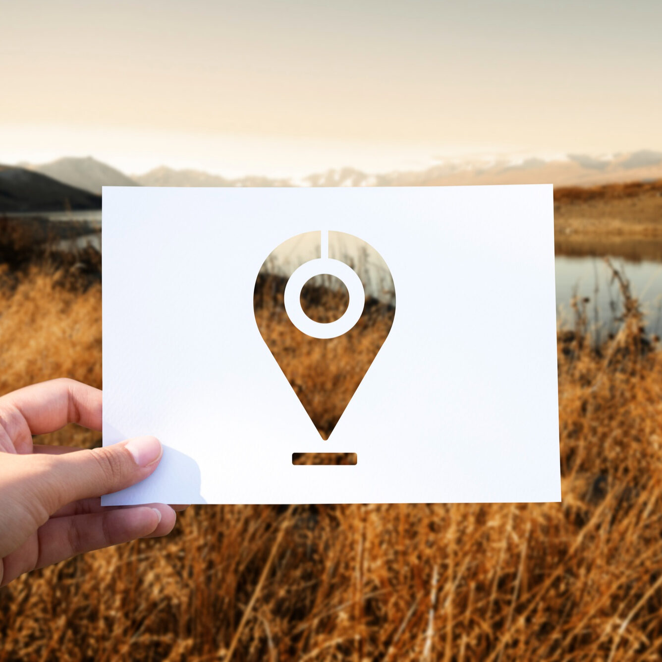 Bild von rawpixel.com auf Freepik Hand hält eine weiße Karte mit ausgestanztem Standort-Symbol vor eine herbstliche Landschaft mit See.