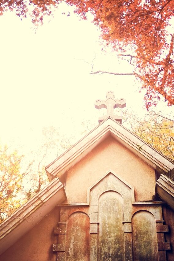 Bild von rawpixel.com auf Freepik Giebelseite eines kleinen Kirchengebäudes mit steinernem Kreuz auf dem Dach. Umgeben von herbstlich gefärbten Bäumen, Licht fällt stark von oben links ins Bild.