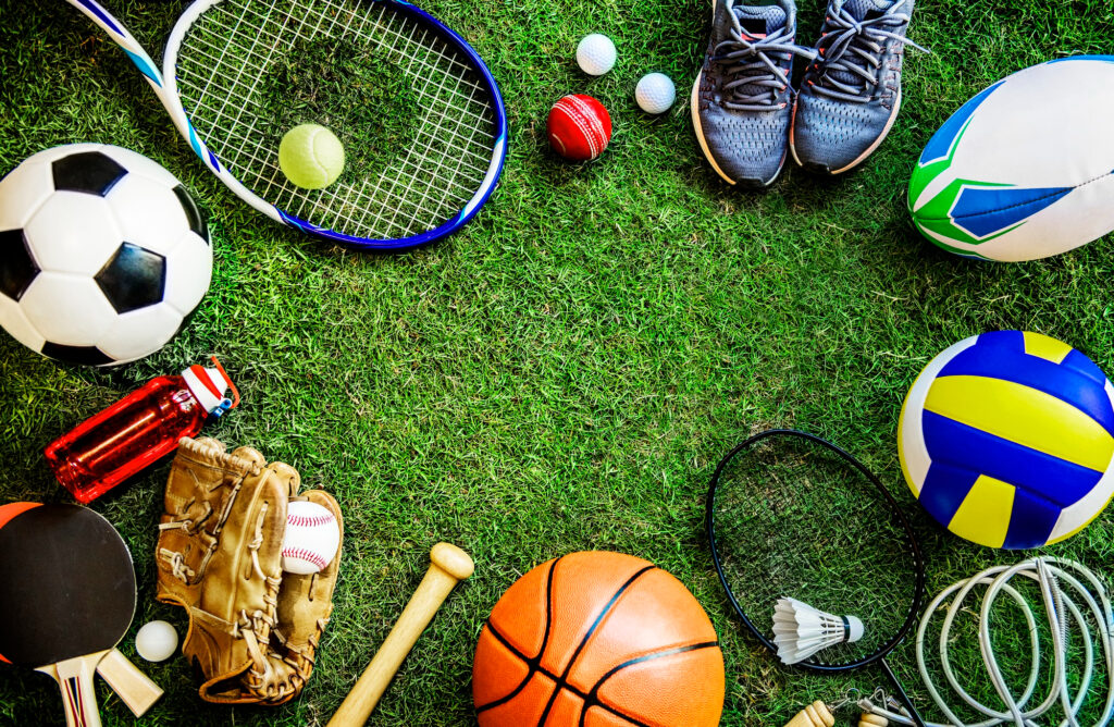 Verschiedene Sportgeräte und Bälle liegen kreisförmig auf Rasen, darunter Fußball, Basketball, Volleyball, Tennis- und Badmintonschläger, Baseballhandschuh mit Ball, Tischtennisschläger, Trinkflasche und Sportschuhe.