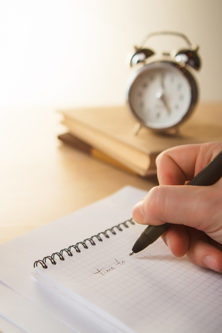 Hand hält einen Stift über einem Notizbuch mit der Aufschrift „time to…“, daneben eine Tasse Kaffee, ein Wecker und Bücher.