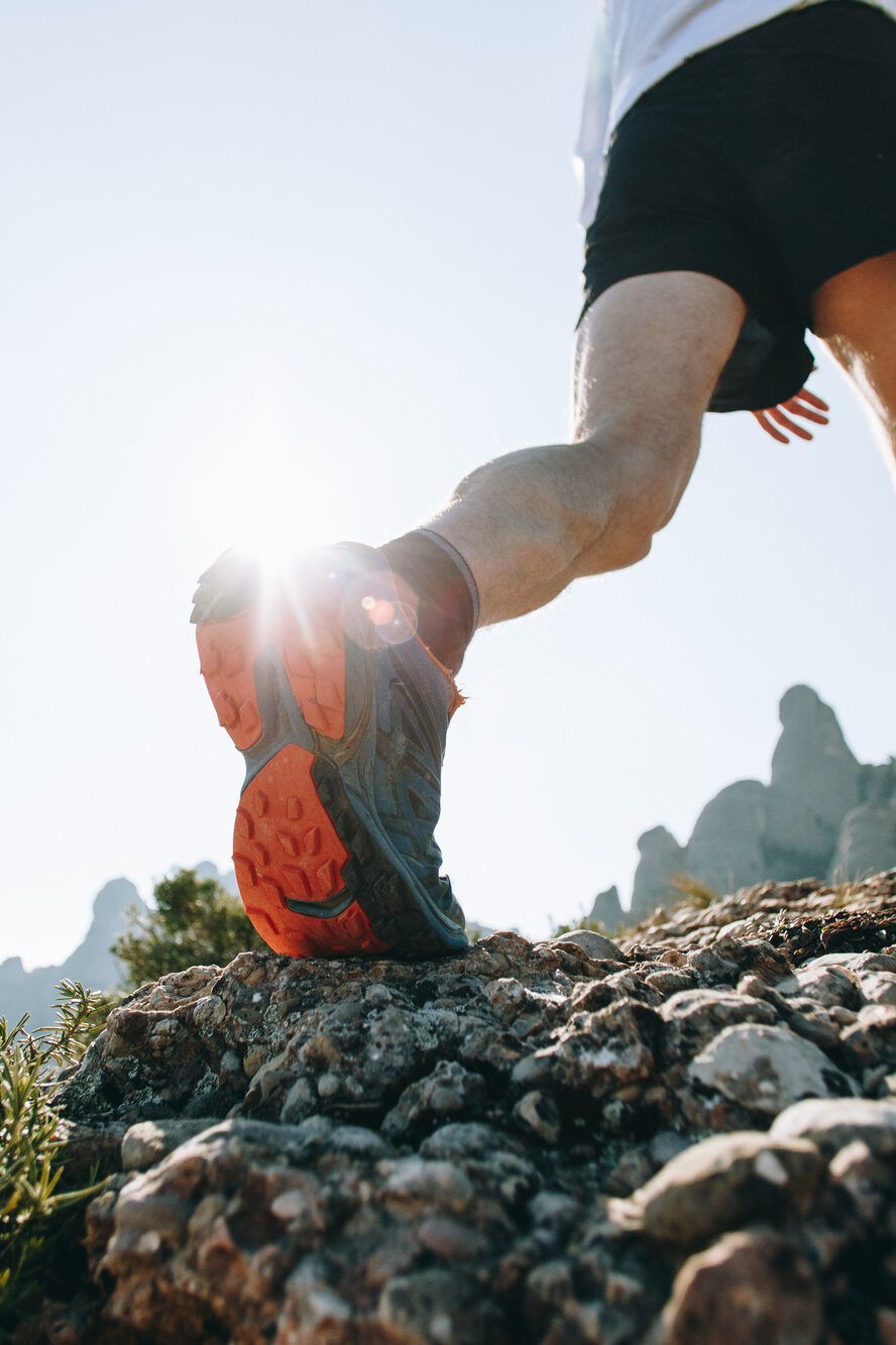 Person in Sportschuhen läuft über felsiges Gelände mit Blick auf Berge und tiefstehende Sonne im Hintergrund.