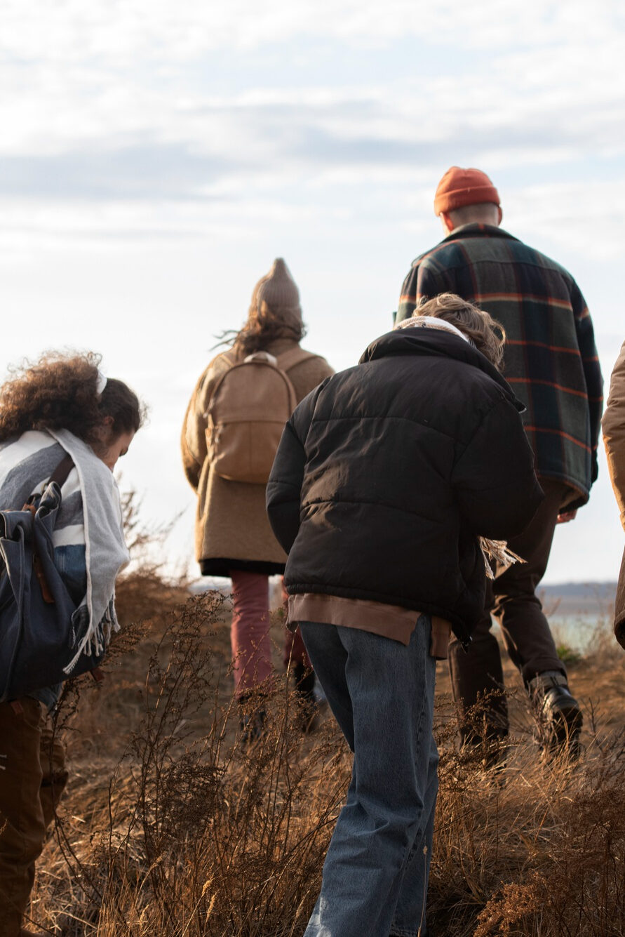 Bild von freepik Sechs Personen gehen mit Rucksäcken und herbstlicher Kleidung einen Hang hinauf. Trockenes Gras am Boden und ein See oder Meer im Hintergrund.