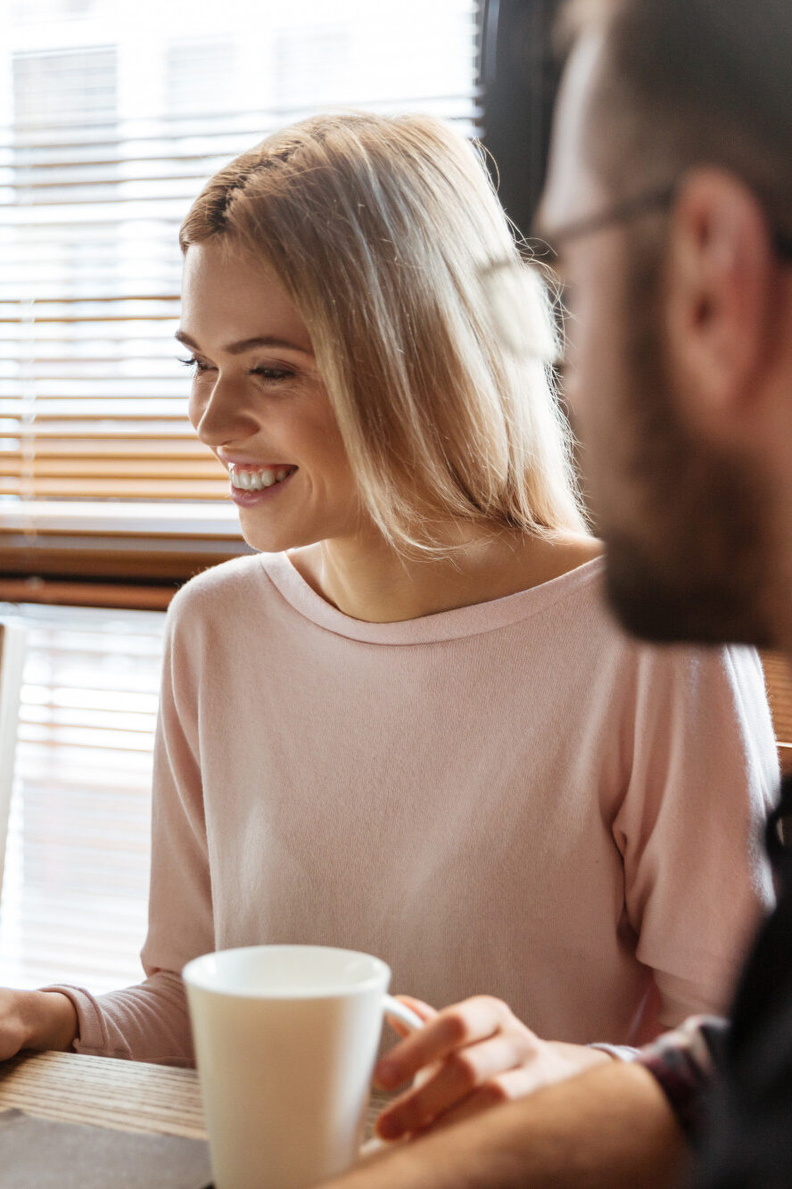 Zwei Personen sitzen an einem Tisch mit Laptop und weißer Tasse. Die Frau in hellem Oberteil lächelt, der Mann trägt eine Brille und blickt auf den Bildschirm.